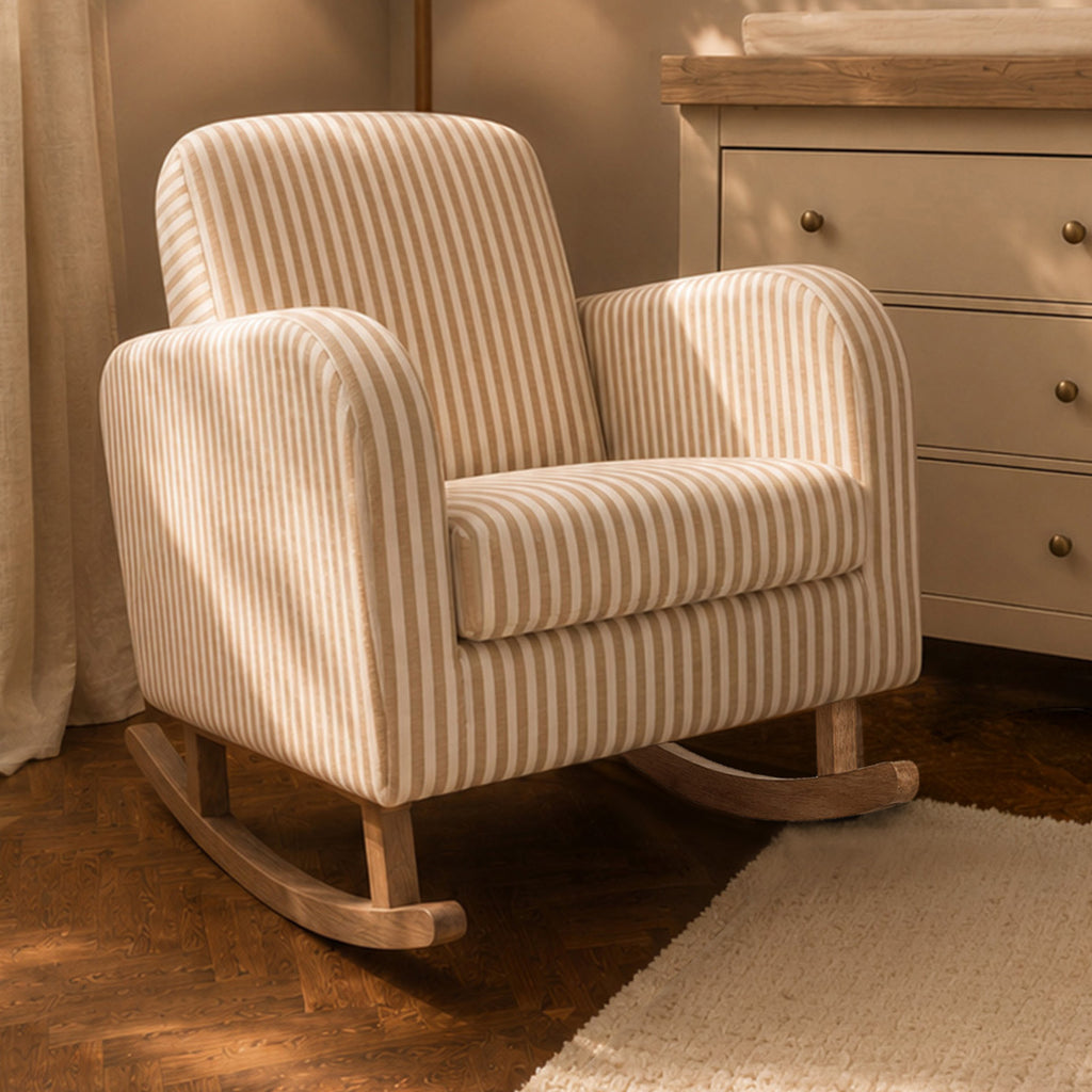 A cream and beige striped fabric rocking chair with a natural wood base, shown in a softly lit nursery or living room setting next to a dresser.