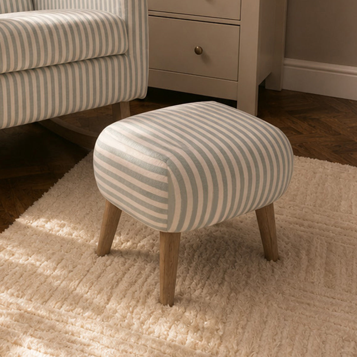 A small upholstered ottoman with cream and blue vertical stripes and light wood legs, shown in a styled room setting next to a matching striped armchair and a cream cabinet.