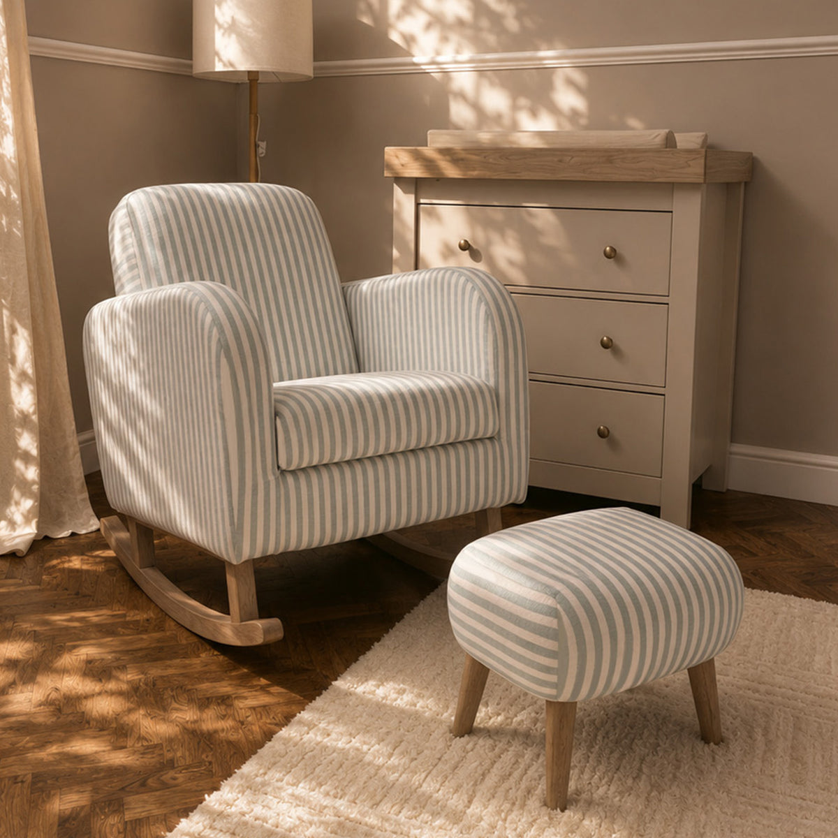 A cream and beige striped rocking armchair with a matching striped ottoman in a styled nursery or living room setting, featuring a wooden dresser, a floor lamp, and warm sunlight filtering through curtains.