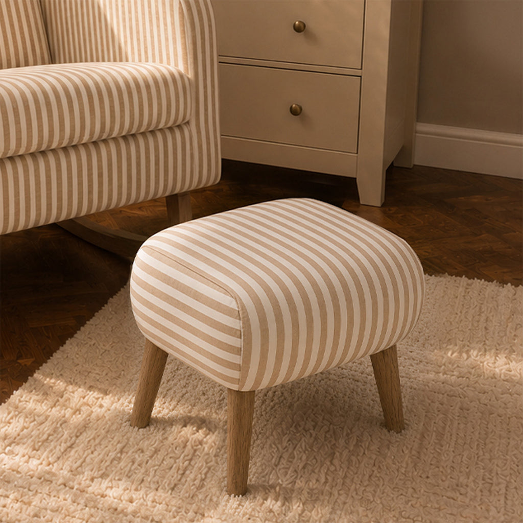 A small upholstered ottoman with cream and beige vertical stripes and light wood legs, shown in a styled room setting next to a matching striped armchair and a cream cabinet.
