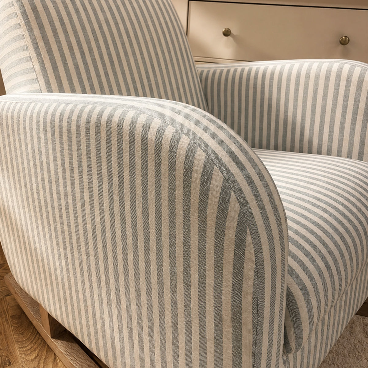 A close-up of a striped upholstered armchair with curved arms and a backrest, shown in a real interior setting with wood flooring and a cream cabinet in the background.