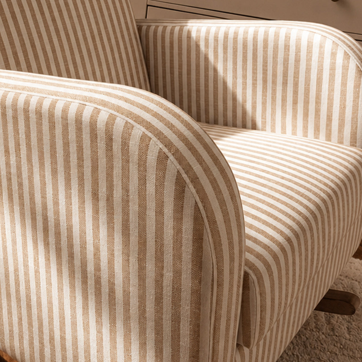 Close-up of upholstered armchair upholstery with narrow vertical beige and off-white stripes, showing the seat, backrest, and curved arm in a softly lit interior.