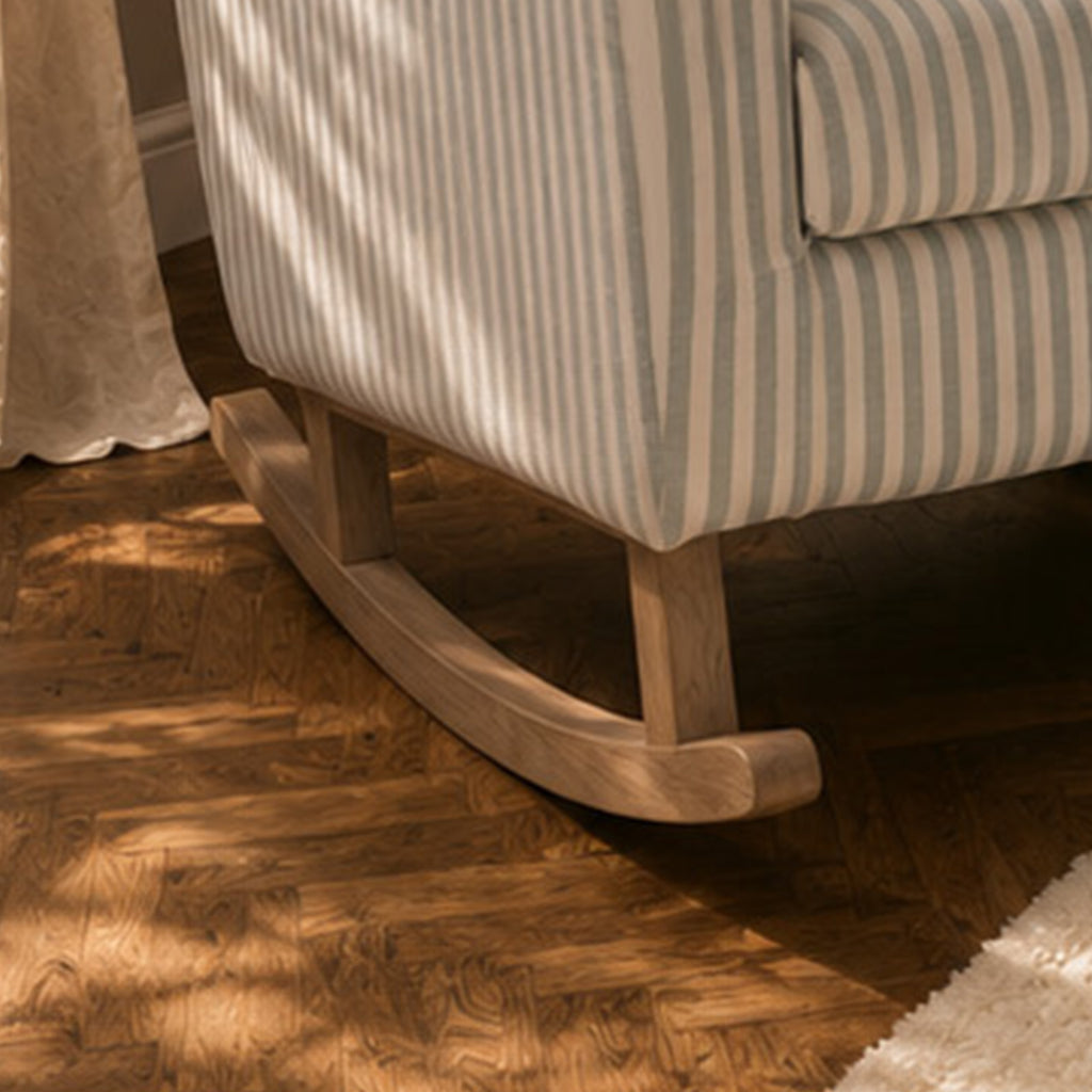 Close-up of a rocking chair with cream and gray vertical stripes and a natural wood rocking base, placed on a dark wood herringbone floor with dappled sunlight.