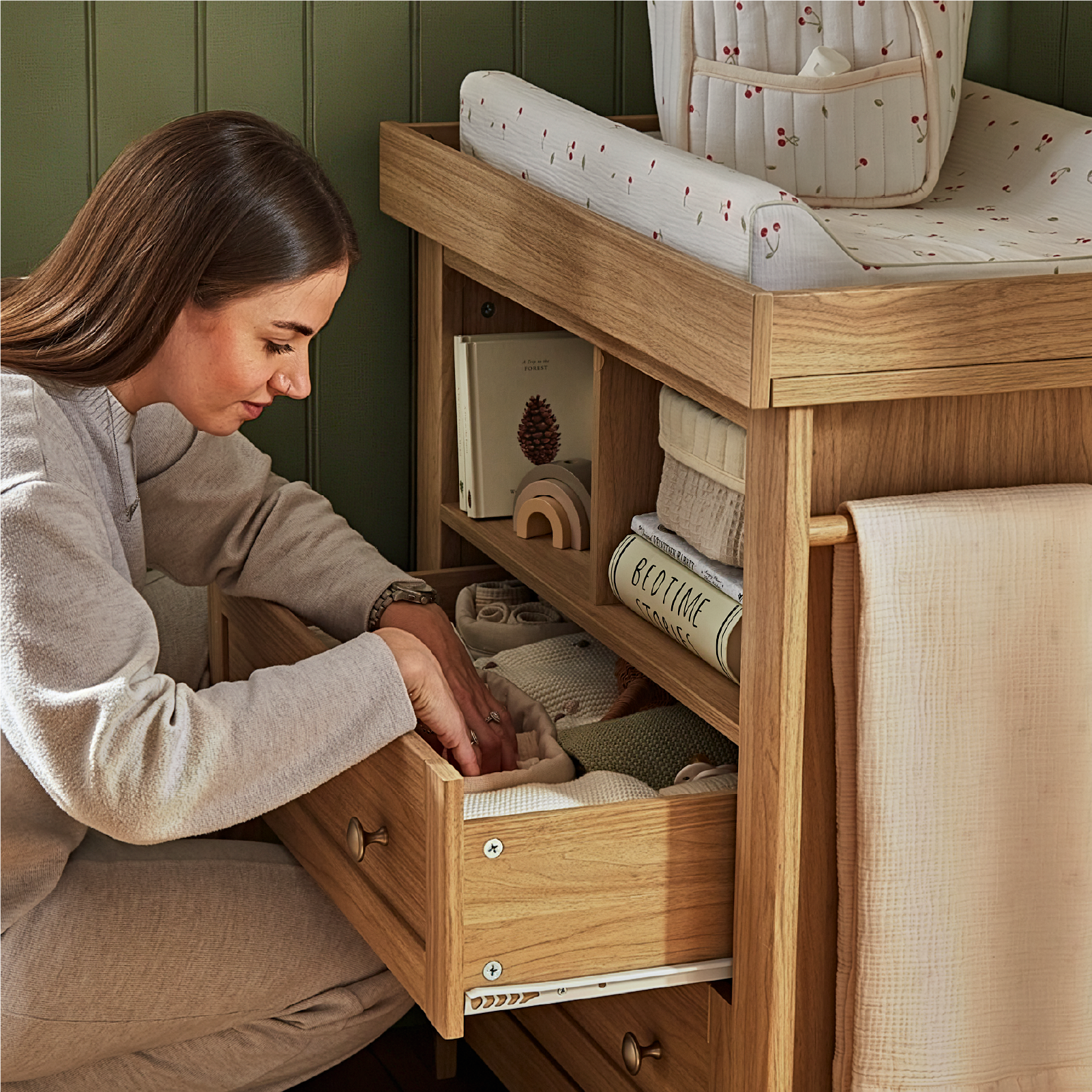 Woman sitting at a wooden style changing table with a drawer open, surrounded by baby items.