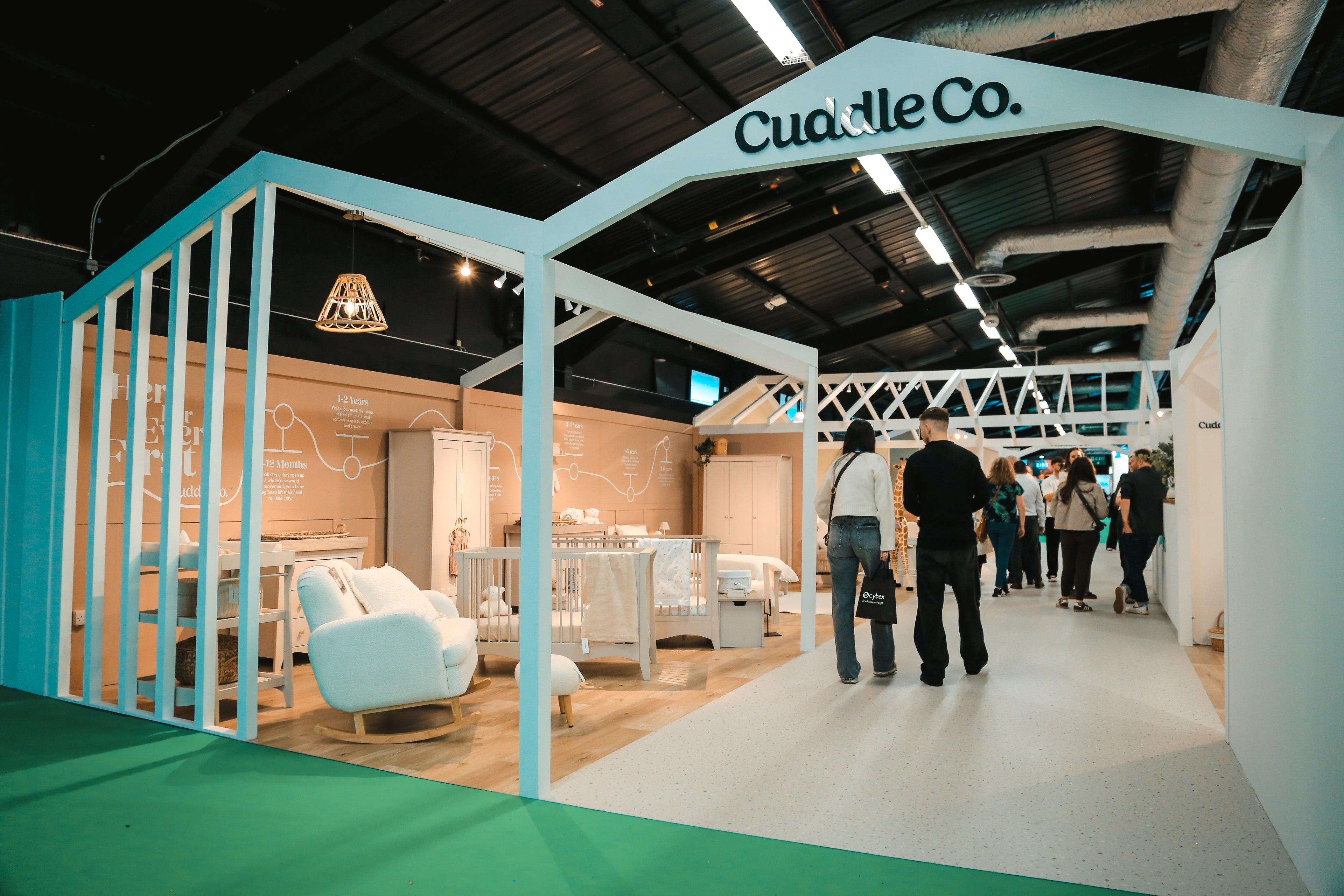 CuddleCo exhibition stand featuring furniture and people walking around.