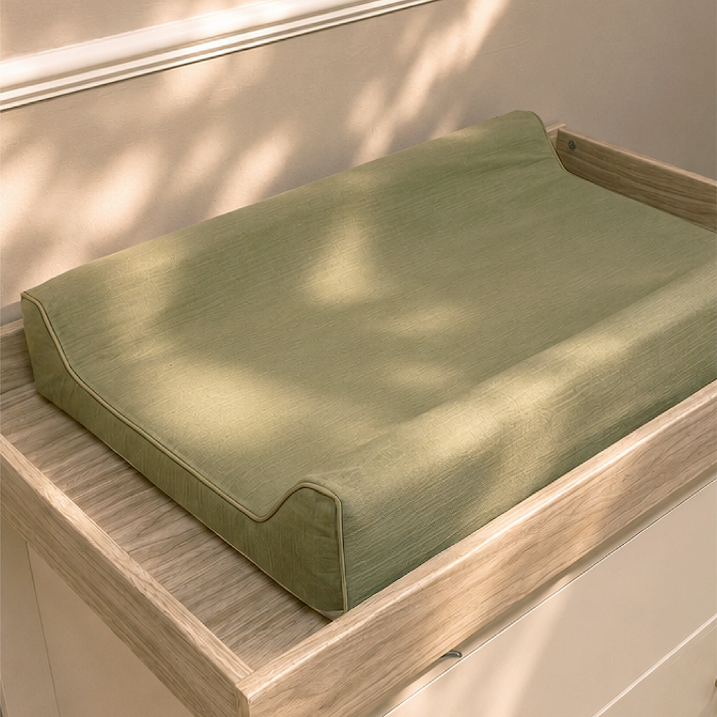An olive green changing pad cushion resting on a light wood changing table dresser, with soft dappled sunlight casting shadows across the surface.