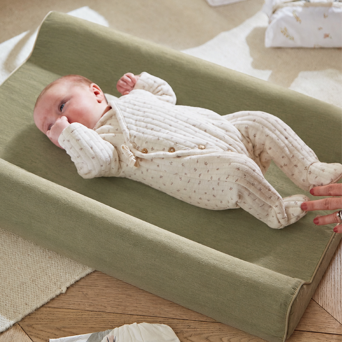 A baby wearing a cream ribbed footed sleeper with tiny gold patterns lies on an olive green changing pad on a wooden floor. A hand is adjusting the pad, and soft home decor items are visible in the background.