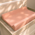 A peach-colored changing pad cushion resting on a light wood changing table, with soft dappled sunlight casting shadows across its surface.