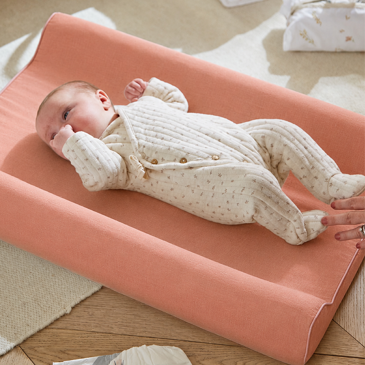 A baby in a cream knit footed romper lies on a peach-colored changing pad on a wooden floor. A hand is adjusting the pad, with a soft home interior background visible.