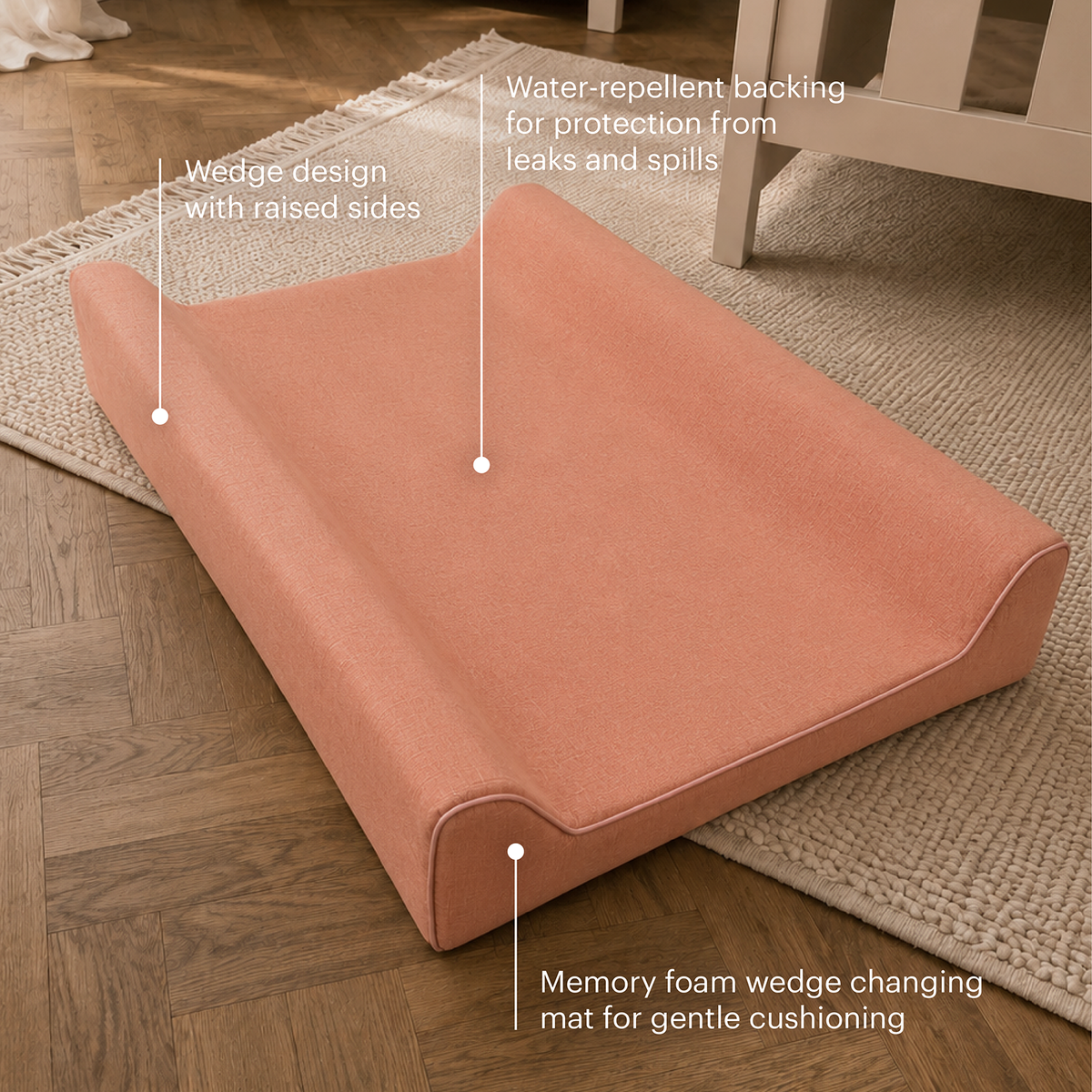 A peach-colored wedge changing mat placed on a wooden floor over a beige rug, with callout text pointing out its raised wedge design, water-repellent backing, and memory foam cushioning.
