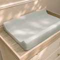A light blue changing pad resting on a light wood changing table dresser with cream drawers.