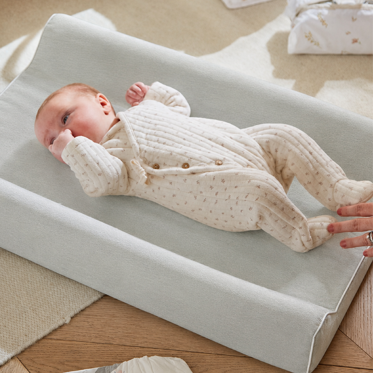 A baby wearing a cream ribbed knit footed sleeper lies on a light blue changing pad on a wooden floor. A hand is adjusting the changing pad, and a patterned blanket is visible in the background.
