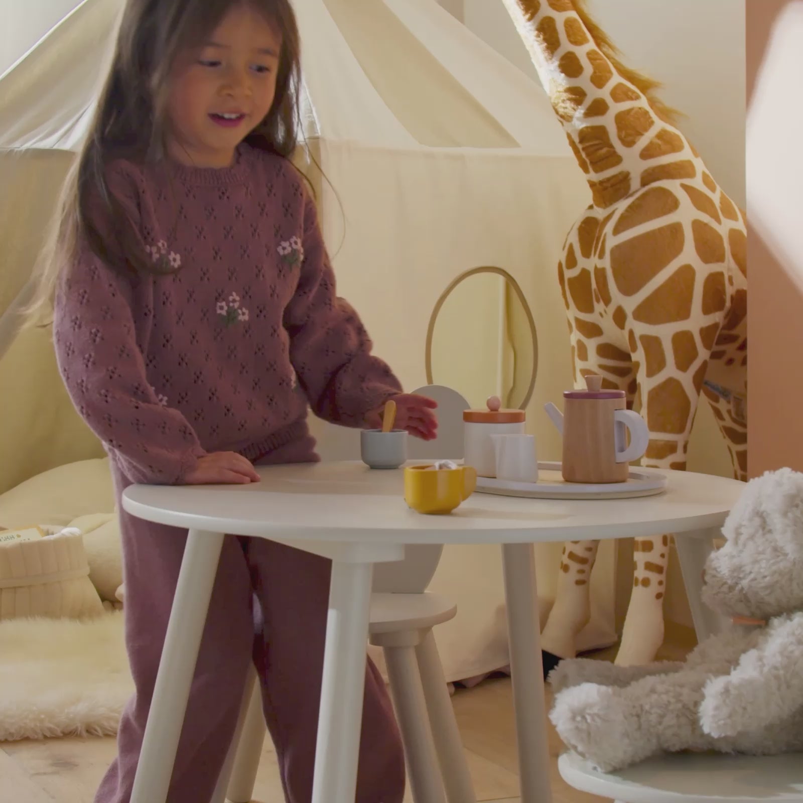 Child and teddy playing with a play house, table and chairs with large toy giraffe in the background